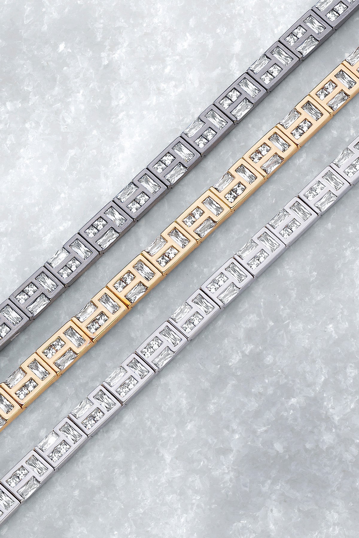 Three APM MONACO labyrinth bracelets in yellow gold, silver, and dark gray on a light iced background.