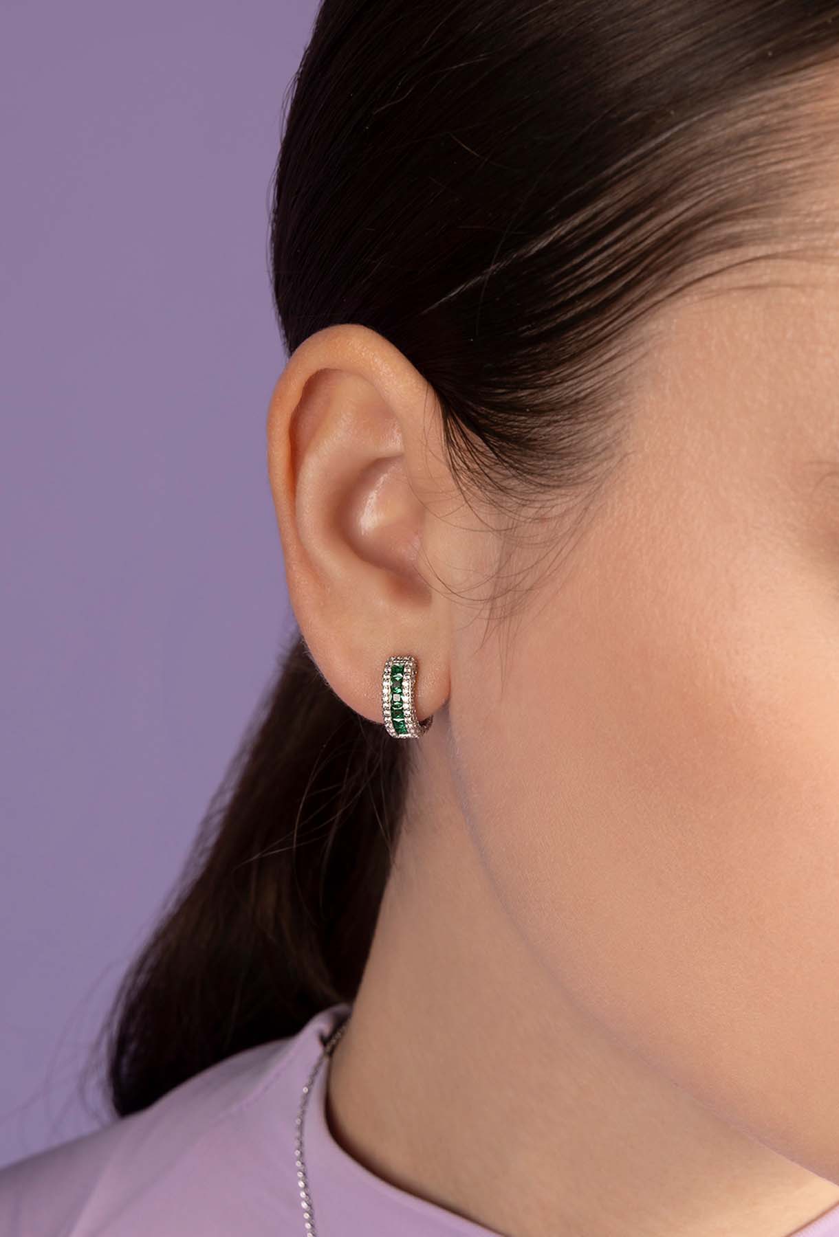 Closeup of an ear wearing APM MONACO green pave huggie earrings with a blurred purple background