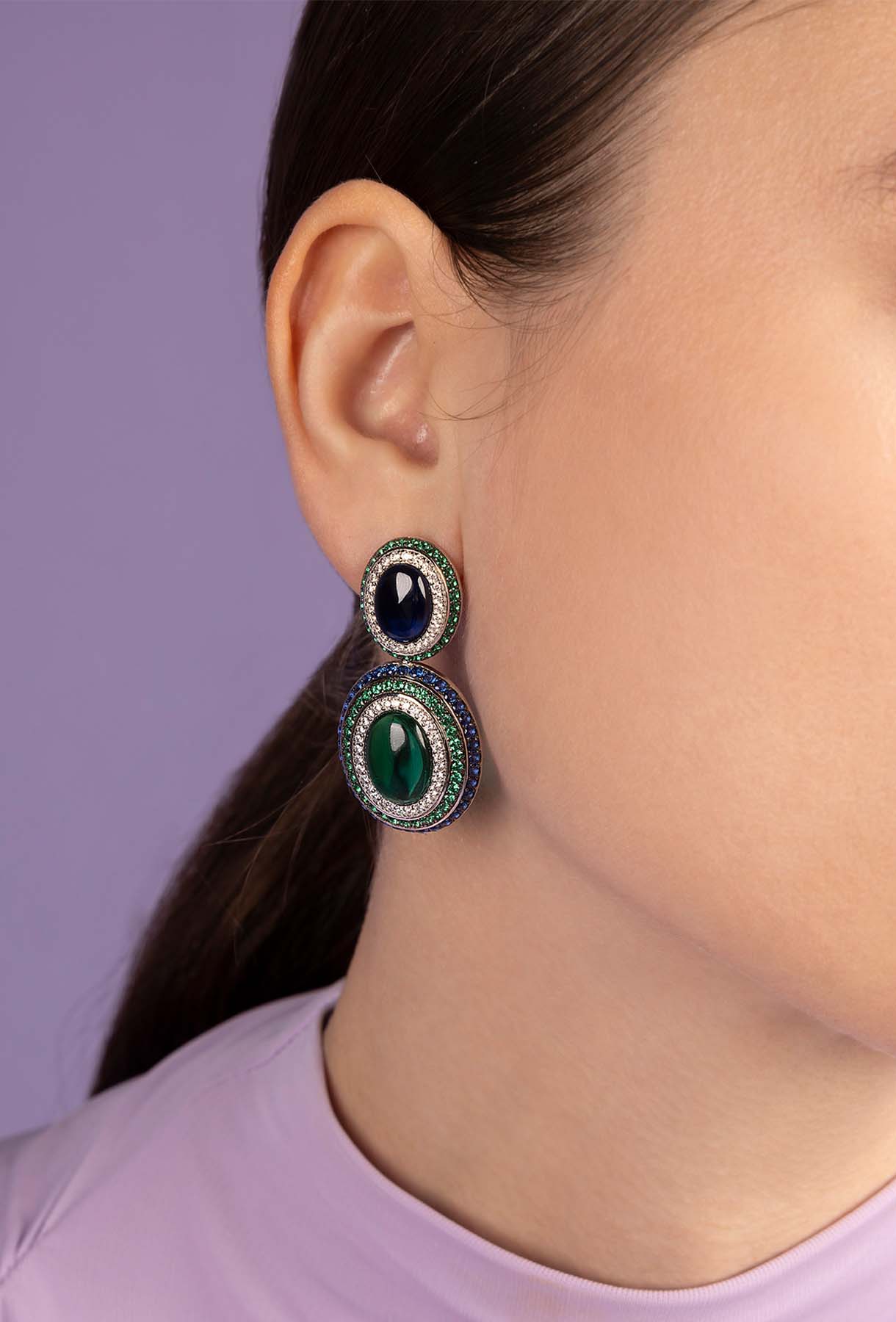 Close-up of a person wearing APM MONACO earrings with green and blue stones against a purple background