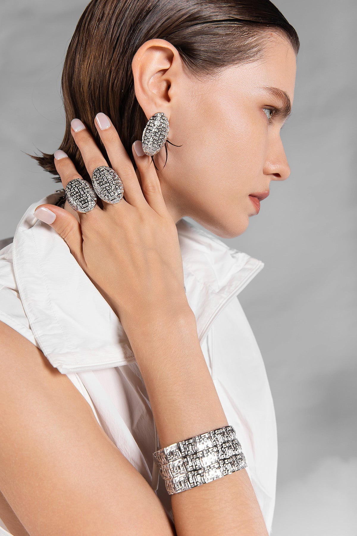 Model wearing APM MONACO jewelry in silver, including rings and an earring on a gray background