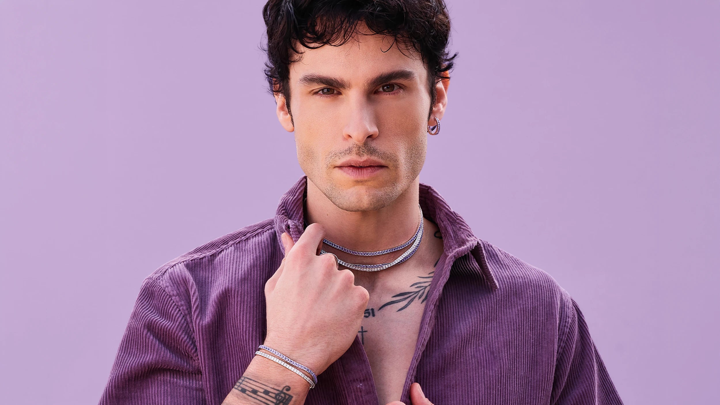  BAPTISTE GIABICONI portant un ensemble de bijoux APM MONACO sur un fond violet clair.