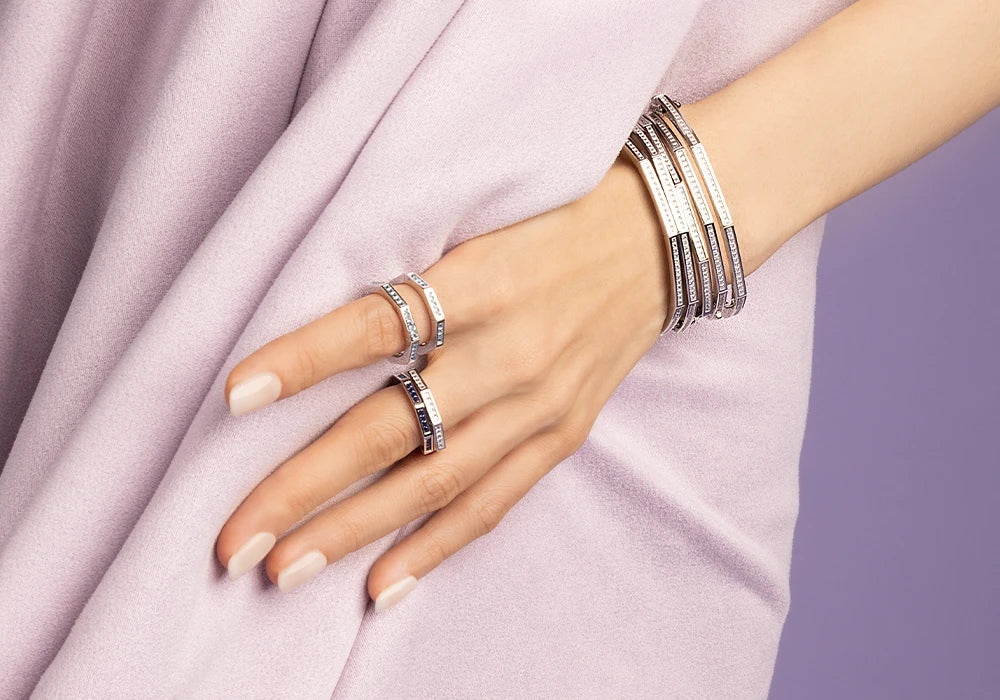 Main portant plusieurs bagues et bracelets en argent de la collection Printemps Lumière d'APM MONACO sur un fond en tissu violet clair.