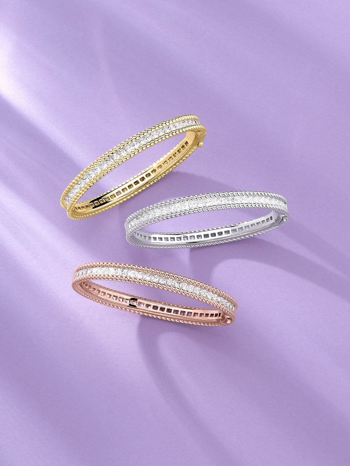 Trois bracelets torsadés APM MONACO en or jaune, en argent et en or rose sur un fond violet clair.